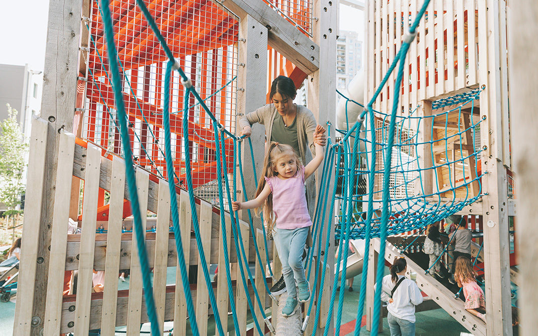 Girl and her mom cross giant rope bridge between timber tower at inclusive playground at Smithe and Richards